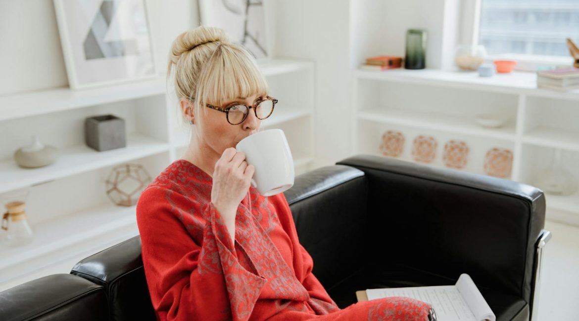 A woman in a red bathrobe drinks coffee while sitting on a sofa indoors