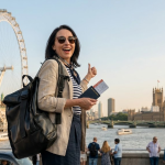 Tourist in London posing with iconic landmarks London Eye and Big Ben in background, holding a passport and giving a thumbs up